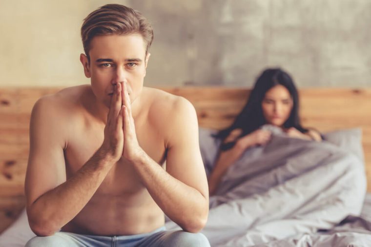 Young couple having a problem. Handsome man is holding hands together while sitting on bed, woman is lying in the background