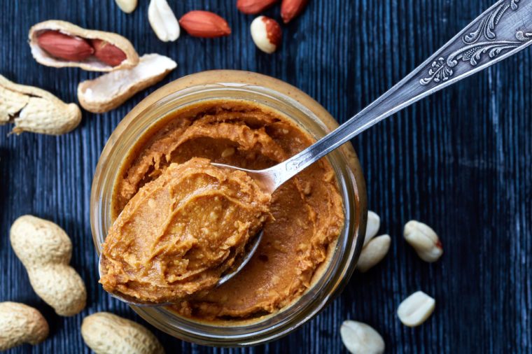 Jar and spoon of peanut butter and peanuts on dark wooden background from top view