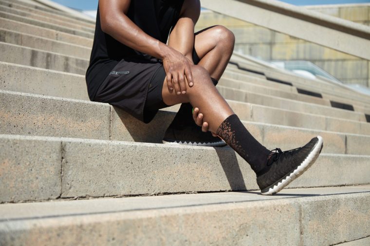 Sports injury concept. Cropped portrait of black male runner wearing black training outfit touching his leg in pain with clasped hands, having sprain or twitch in his knee after running exercises