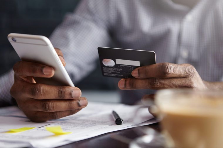 Cropped shot of African-American businessman paying with credit card online making orders via Internet. Successful black male holding plastic card making transaction using mobile banking application