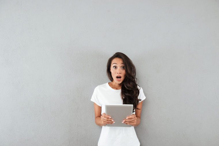 Shocked casual asian woman holding pc tablet and looking at camera while standing isolated over gray background