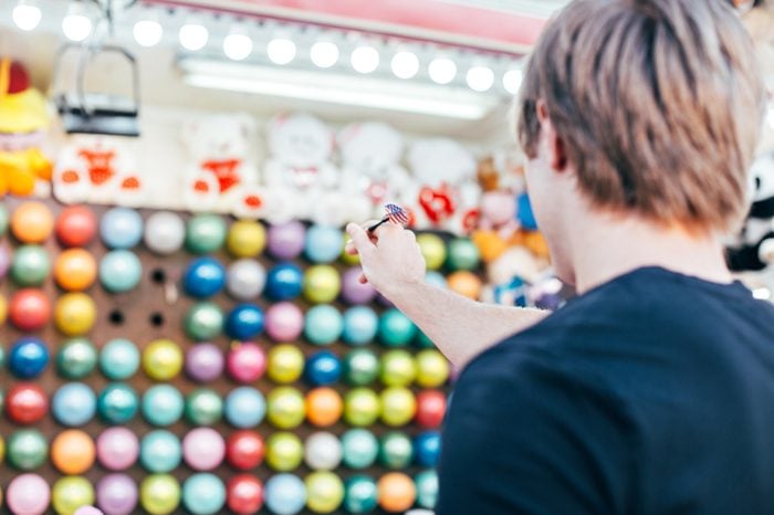 Young man aims at wall of colourful air balloons in order to win teddy bear or plush toy prize for his girlfriend, during romantic date at amusement park or carnival