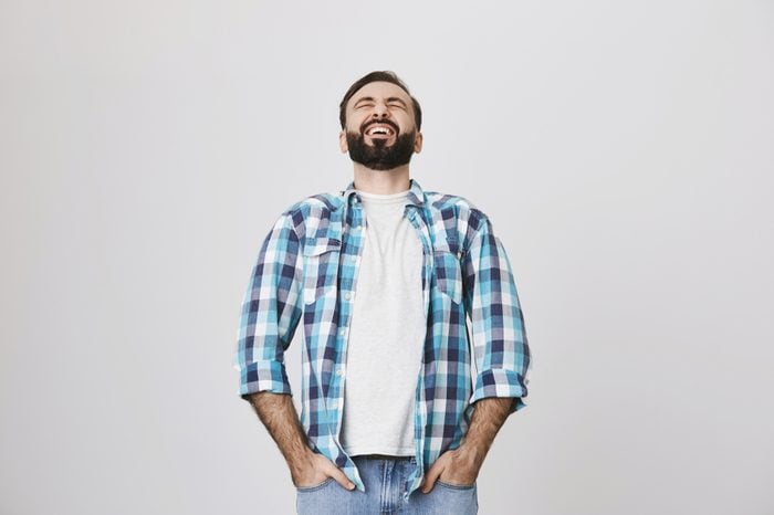 Studio shot of handsome european adult with beard and moustache, laughing out loud, stooped back, holding hands in pockets of jeans, over gray background. Guy watches stand up show on TV