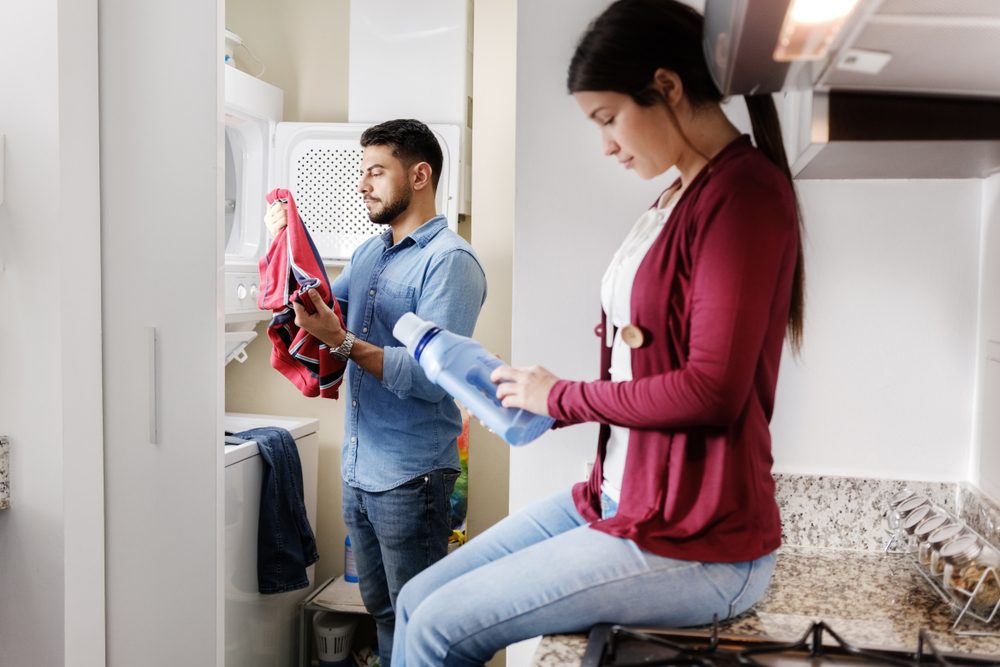 Young couple doing housework and chores. Man putting clothes inside washing machine and girlfriend sitting on kitchen counter, reading label on cleansing box.