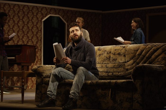 Medium shot of an actor reciting his lines on a sofa while other actors rehearsing in the background