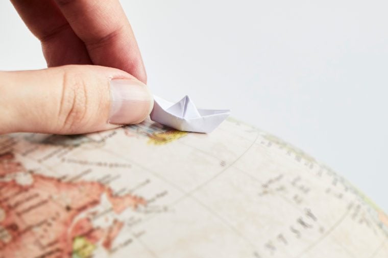 Closeup of a hand holding a little paper boat on a globe on white background.