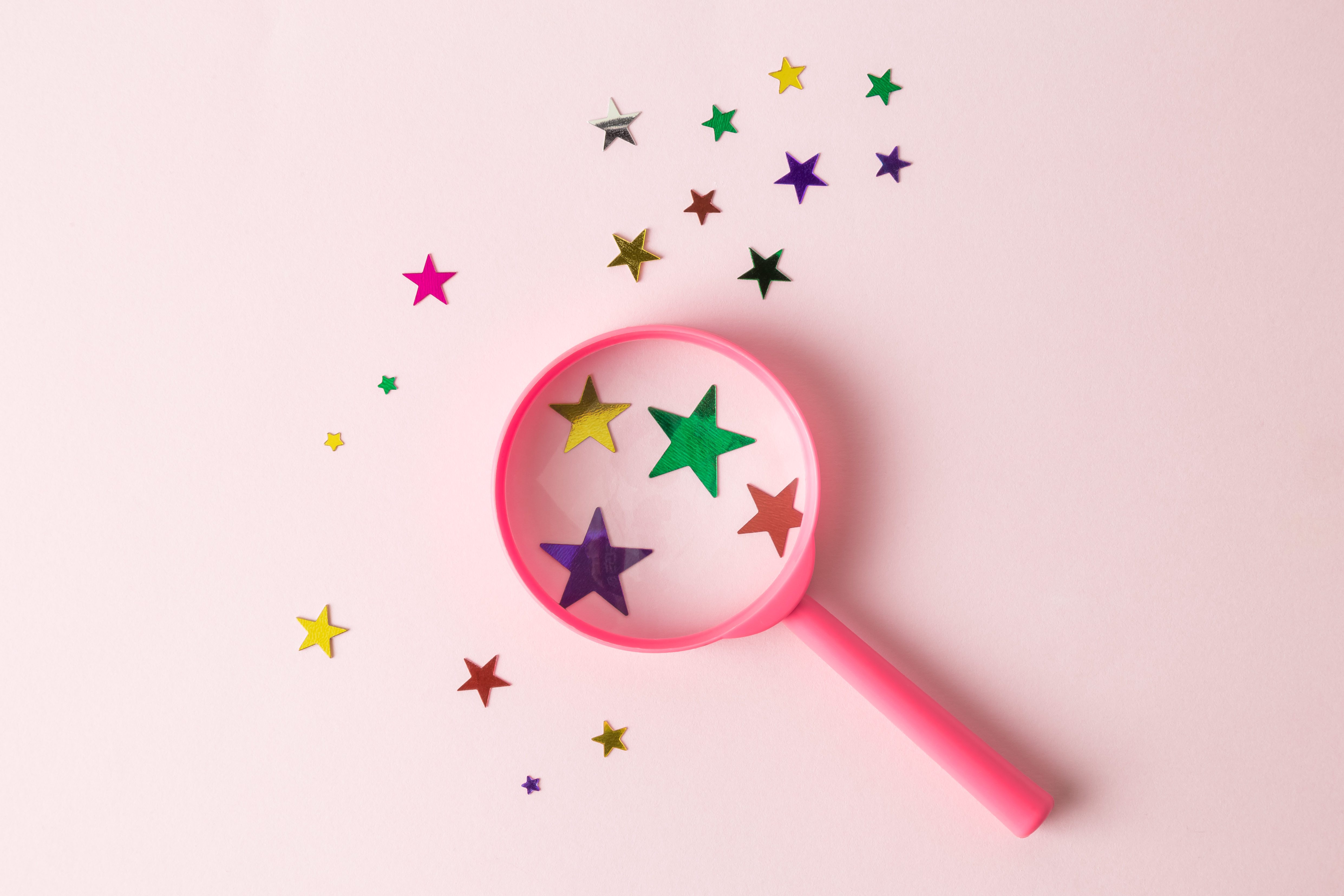 Top view of magnifier and different sizes multicolored stars on pastel rose background abstract.