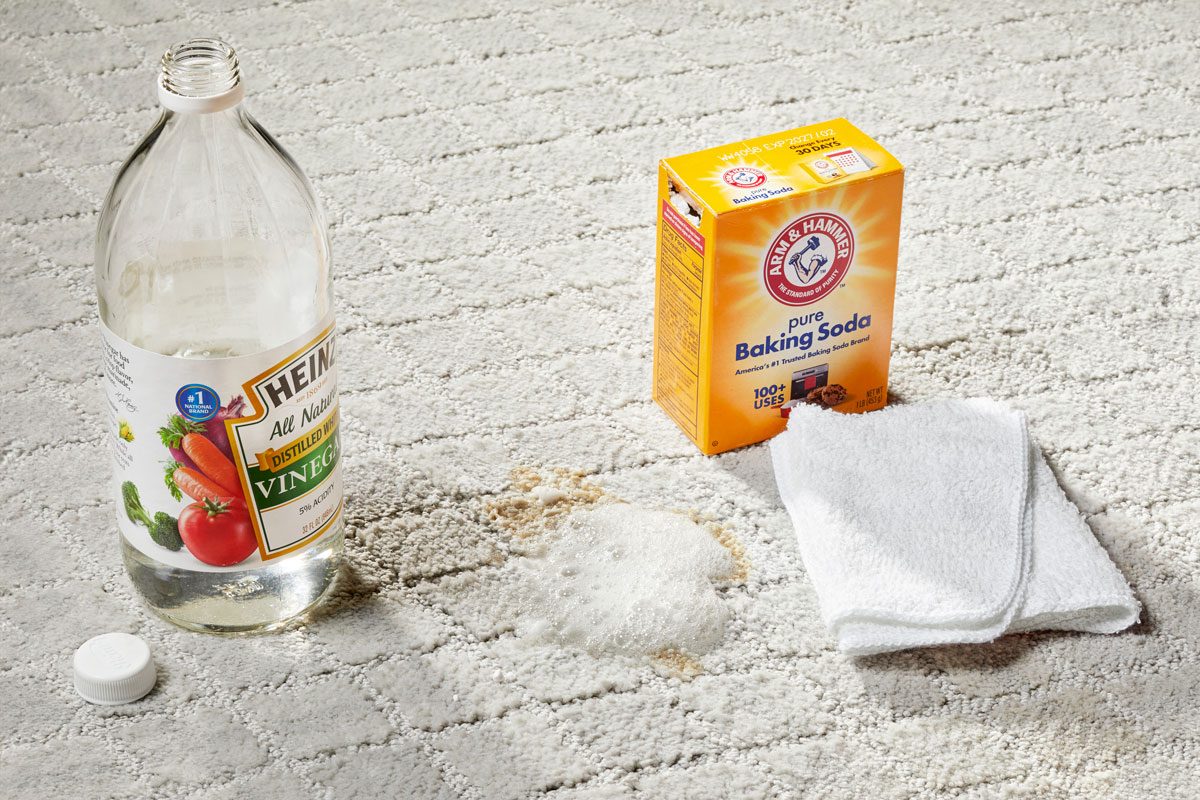 Baking soda and vinegar solution bubbling on a stain on a light gray carpet with vinegar bottle, baking soda box, and cleaning cloth nearby for Homemade Baking Soda Carpet Cleaner