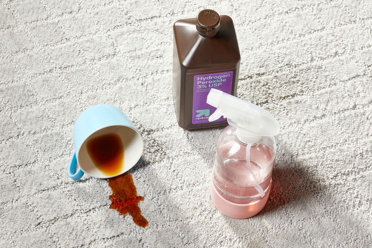 spilled coffee cup and coffee stain on a light gray carpet next to a bottle of hydrogen peroxide and a spray bottle for Homemade Hydrogen Peroxide Carpet Cleaner