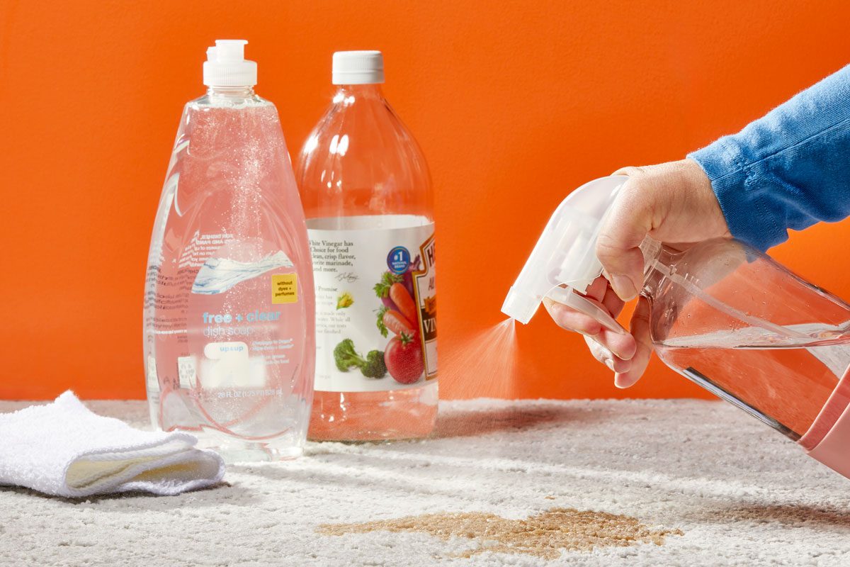 Hand using a spray bottle on a stain with dish soap, vinegar, and cleaning cloths nearby in the background for Homemade Vinegar Carpet Cleaner