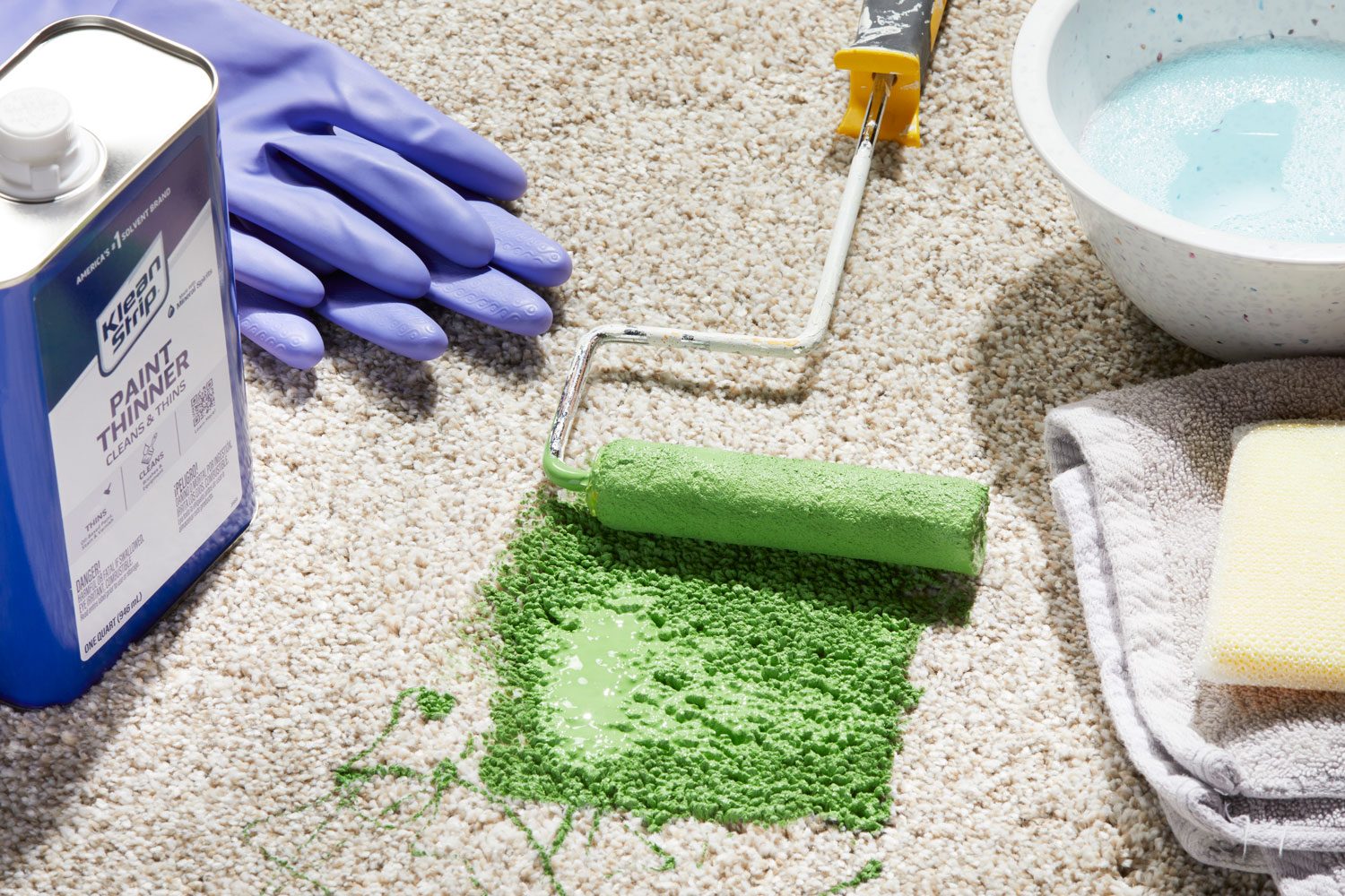 green oil-based paint and paint roller on a light carpet causing a stain; cleaning products surrounding: paint thinner, gloves, bowl of soapy water, towel, sponge