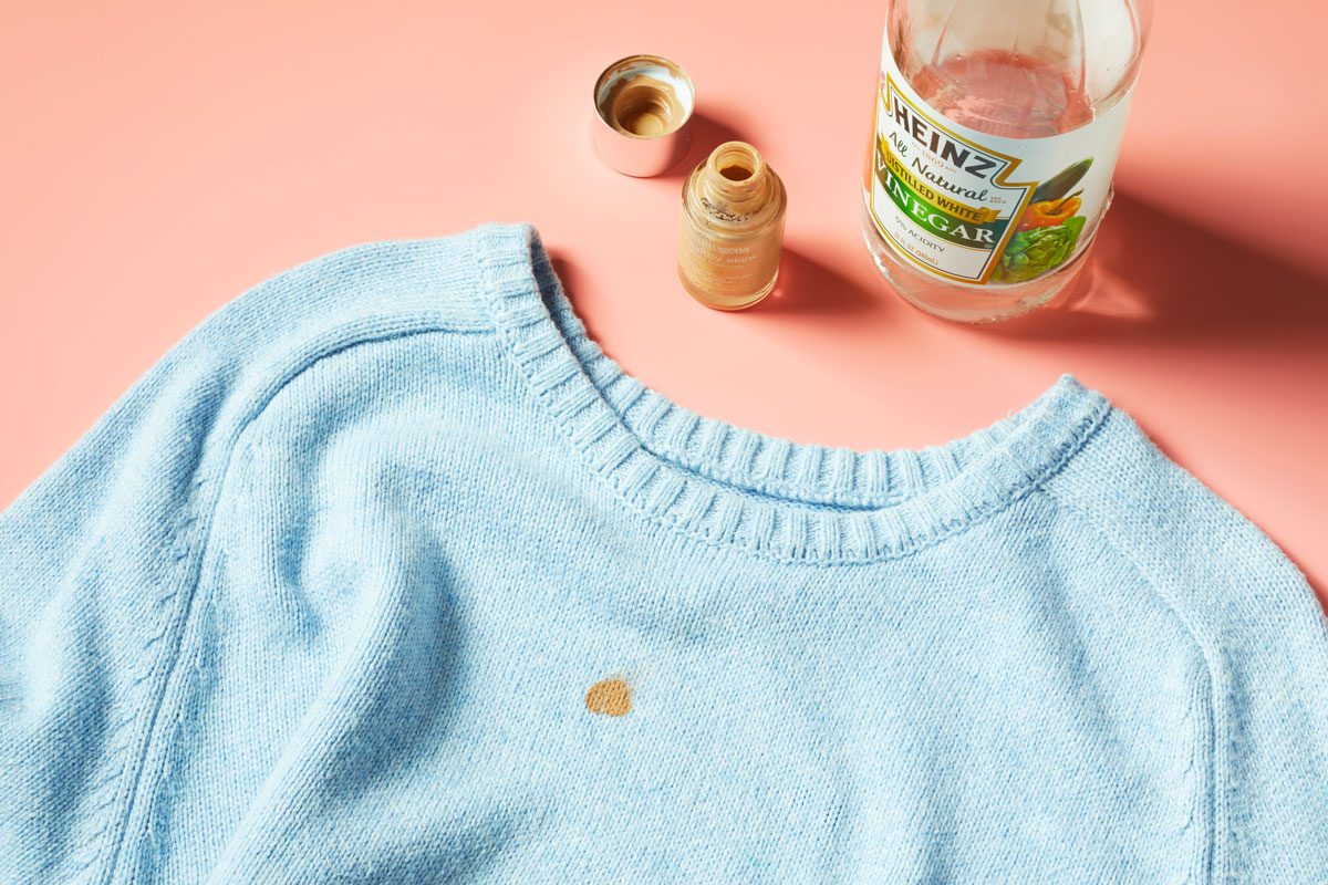 Liquid foundation stain on a blue sweater with liquid foundation makeup and bottle of vinegar nearby. peach background.