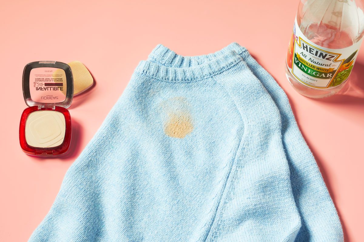 powder foundation stain on a blue sweater with powder foundation makeup and bottle of vinegar nearby. peach background.