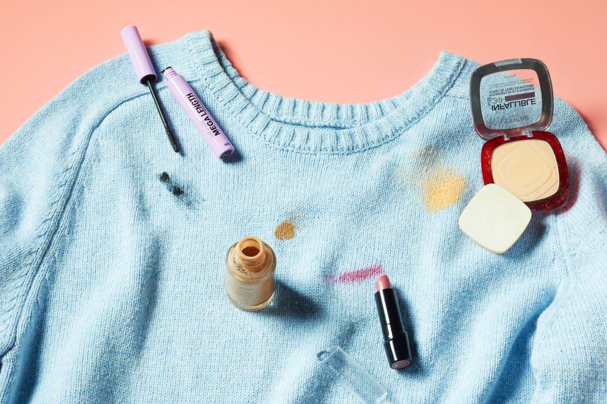 a blue sweater with makeup stains on a peach colored background. the sweater has a mascara stain, liquid foundation stain, lipstick stain, and powder foundation stain.