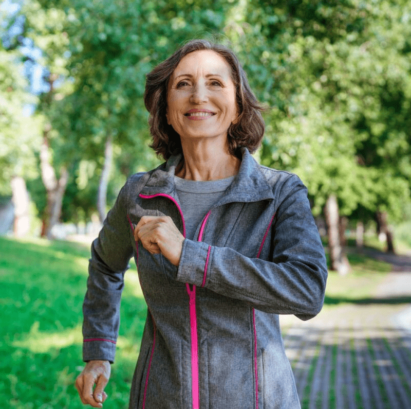 smiling woman in gray jacket strolling briskly in lush green park