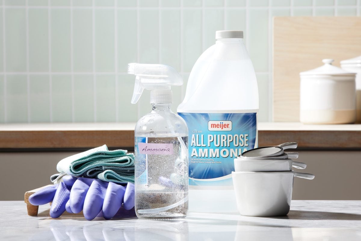 bottle of ammonia on a kitchen countertop surround by a spray bottle, measuring cups, cleaning cloths, and rubber cleaning gloves