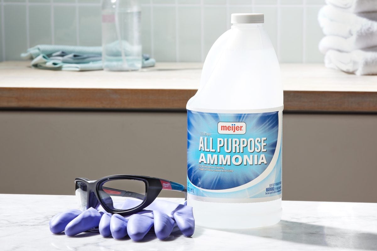 bottle of ammonia next to rubber gloves and eye protection glasses on a marble countertop. In the background, a spray bottle, cleaning cloths, and white towels are visible