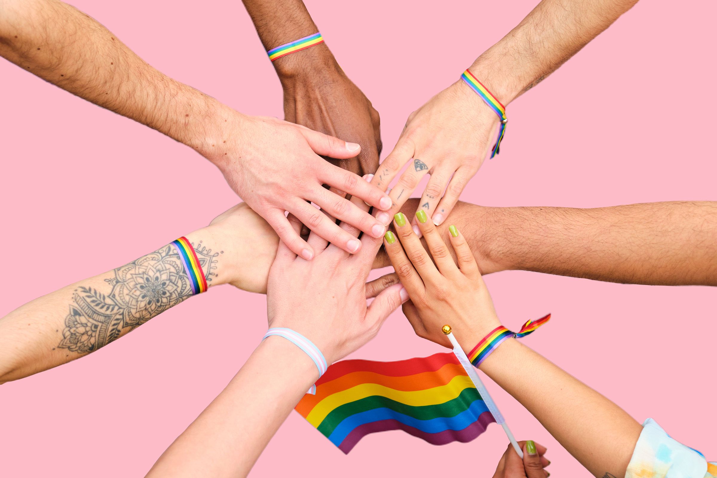 A group of people stacking hands and holding rainbow flags on a pink background