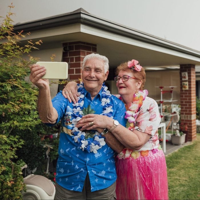 An elderly couple dressed in colorful Hawaiian attire takes a selfie outside their home. The man wears a blue Hawaiian shirt and a floral lei, while the woman wears a floral lei and a pink skirt with a flower in her hair. Both are smiling and appear happy.