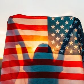 Independence day. Silhouette of woman holds an American flag in her hands, raised above her head. Back view. In the background, sunset and mountains. The concept of American National Holidays