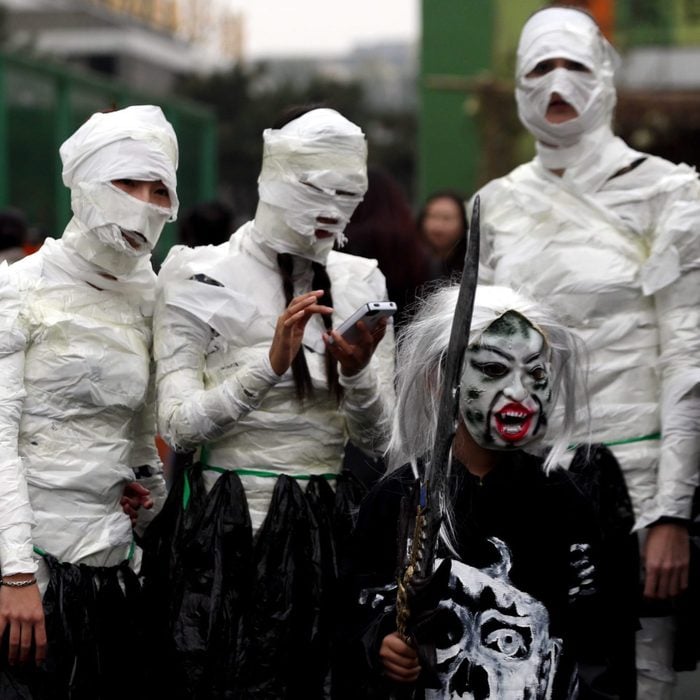 Four people dressed in mummy costumes, with their faces and heads wrapped in white bandages, stand together. Another person, dressed in a ghoulish outfit with white hair and face paint, holds a sword. They appear to be in a festive or Halloween-themed setting.