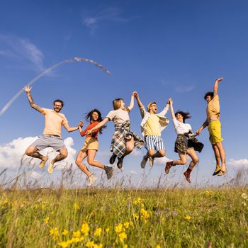 Low Angle View Of Group Of Happy Friends Having Fun While Holding Hands And Jumping In Meadow