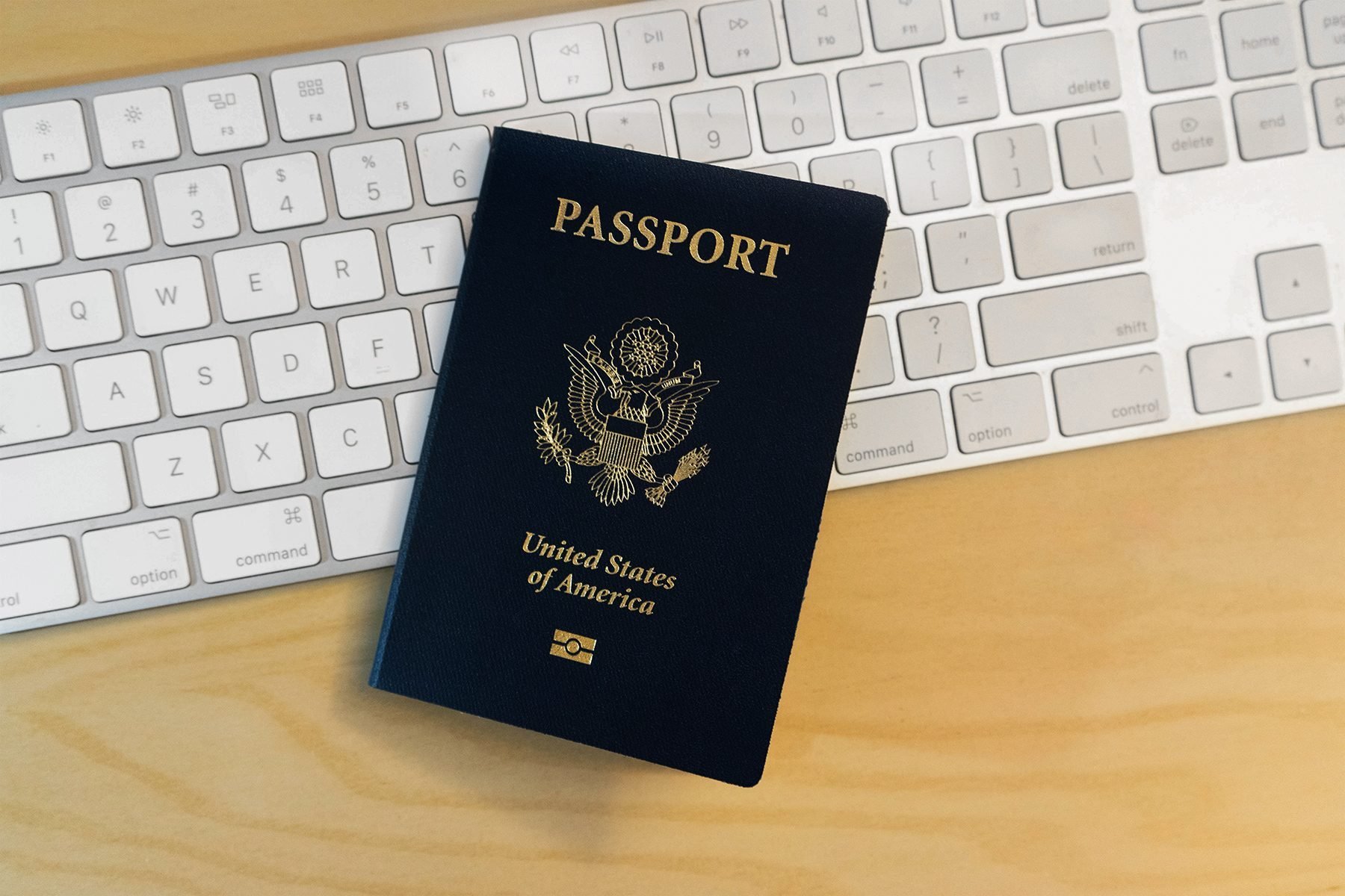 A United States passport lies on a wooden desk, partially covering a white computer keyboard. The passport is closed and its cover, displaying the U.S. emblem and the word "PASSPORT," is clearly visible.