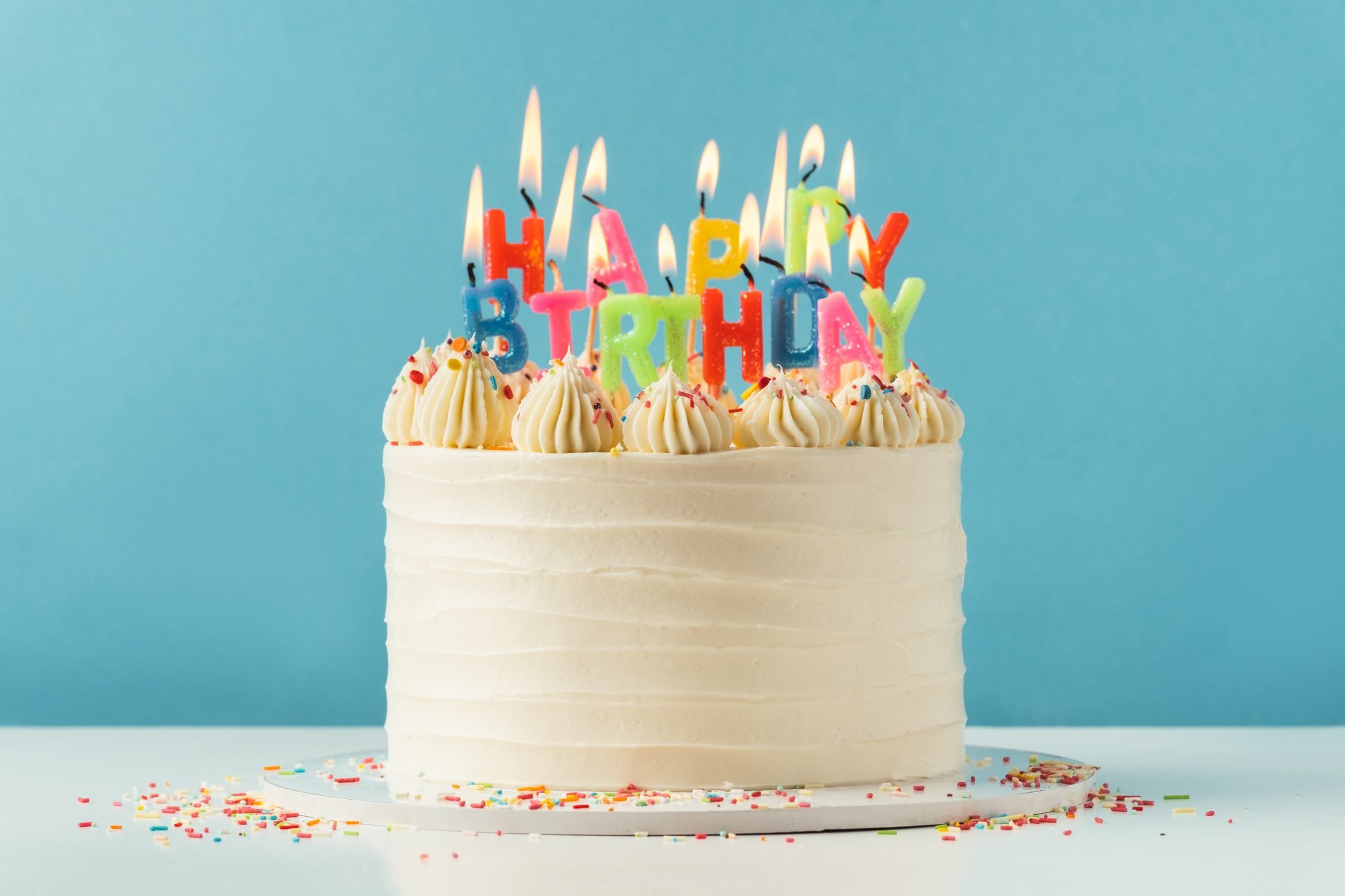 Birthday cake with white cream cheese frosting decorated with multicolored lit happy birthday text shaped candles on a blue background. Tradition of making a wish while blowing out candles on a cake