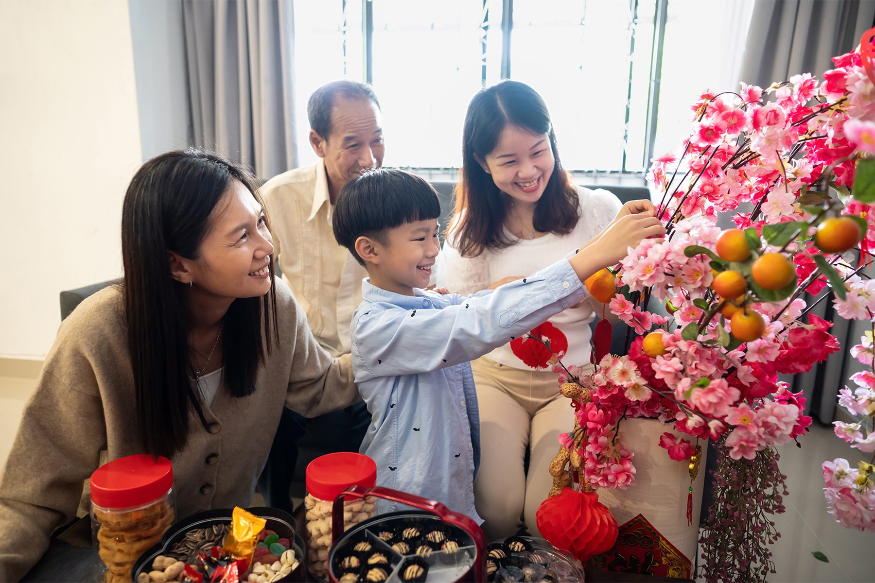 A family of four, including an older man, two women, and a child, decorates a vibrant tree with pink blossoms and red ornaments indoors. They are smiling and appear happy, with traditional snacks on the table in front of them.