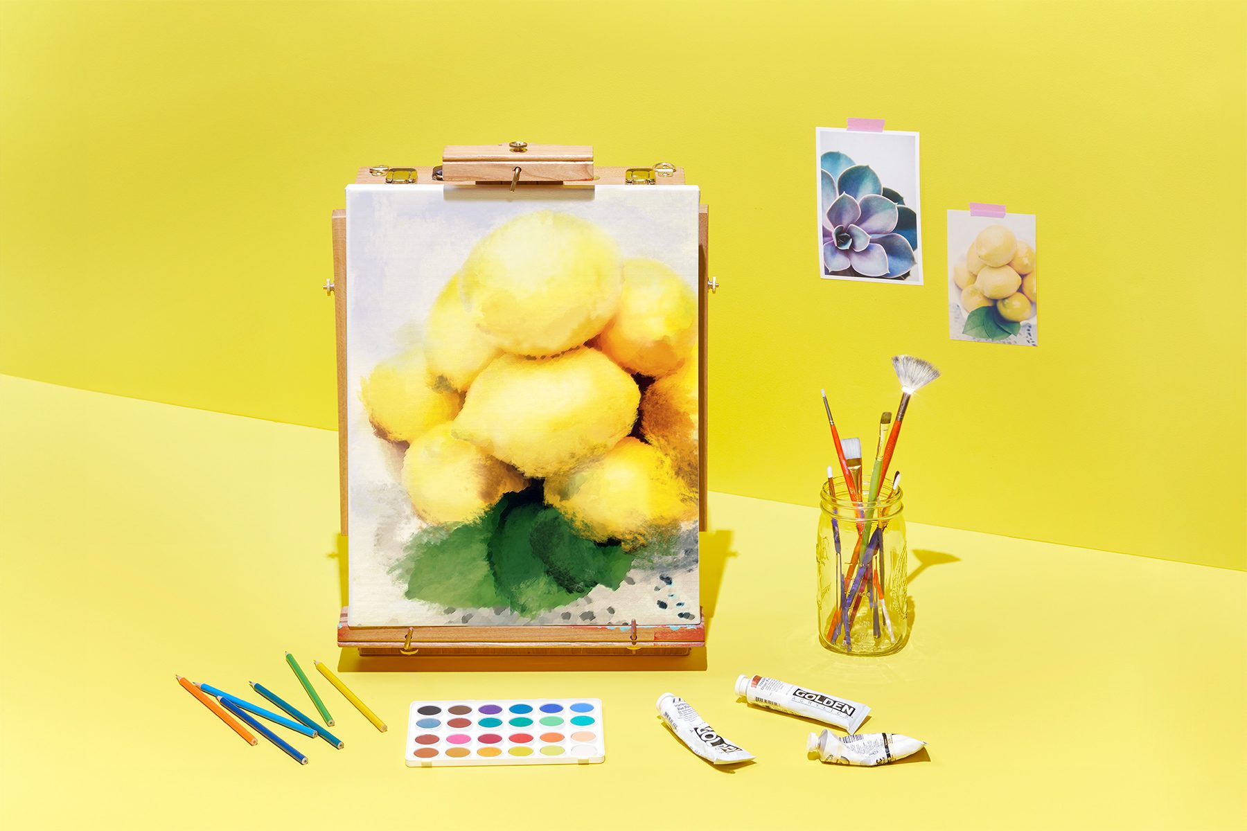 A painting of yellow lemons on an easel sits on a yellow surface with a brighter yellow background. Beside it, there are watercolor paints, colored pencils, paint brushes in a glass jar, and a couple of reference photos of the lemons and a succulent plant.