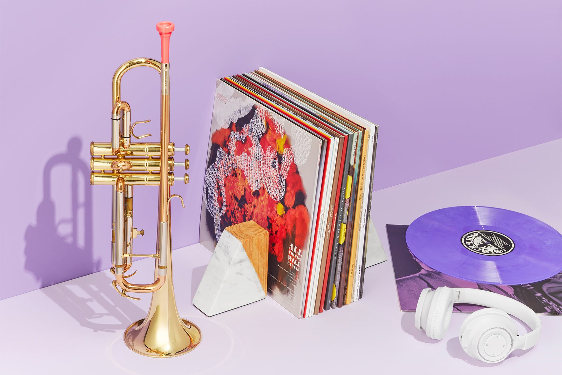 A polished trumpet, several vinyl records propped up between two geometric bookends, a purple vinyl record spinning on a turntable, and a pair of white headphones rest on a light purple surface. The background is a light lavender color.