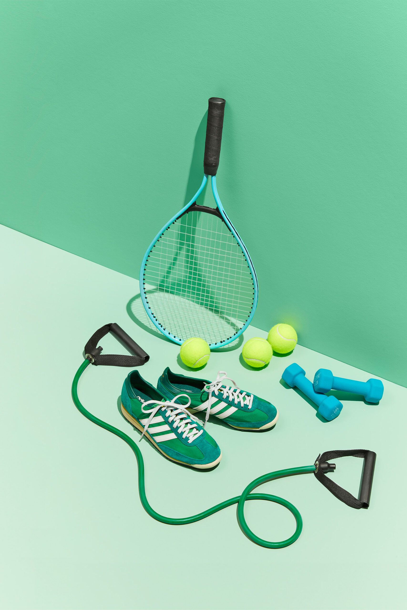 A tennis racket, three yellow tennis balls, two small blue dumbbells, green athletic shoes with white laces and stripes, and a green resistance band are arranged on a light green background.